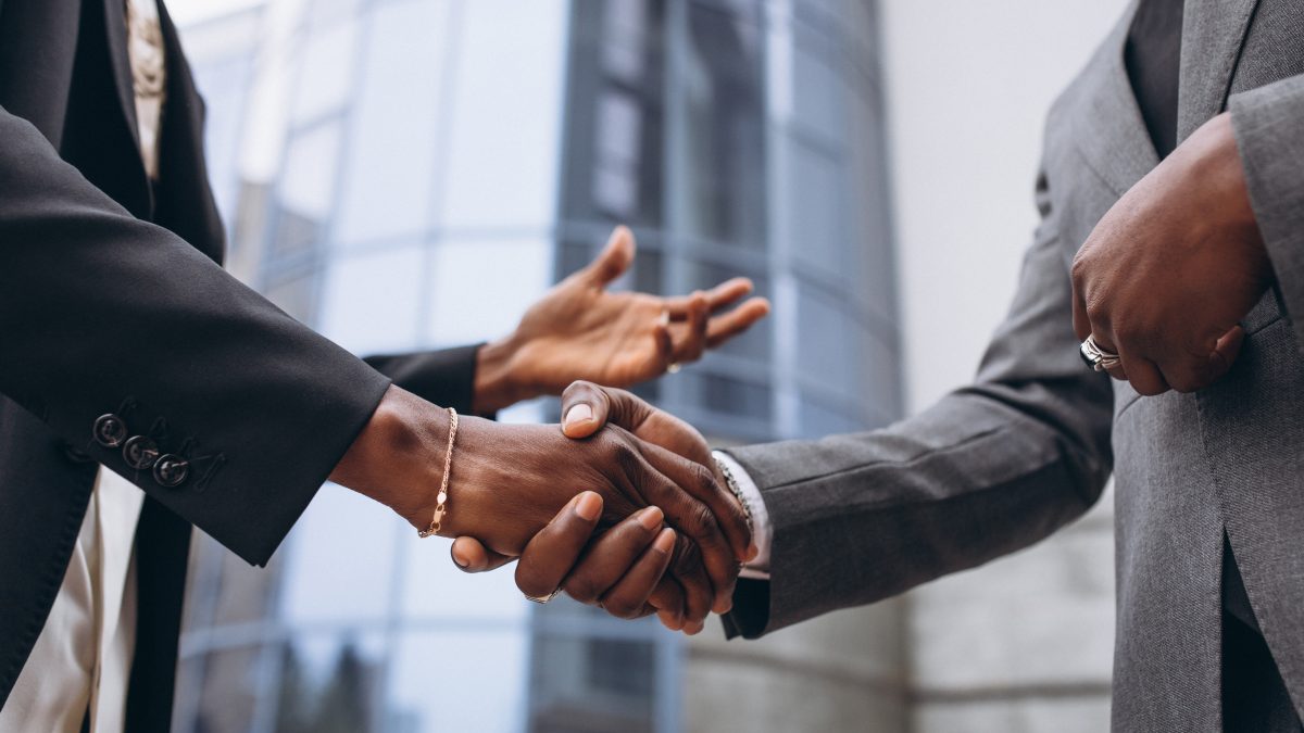 Two business professionals in formal suits shaking hands outdoors in front of a modern office building, symbolizing partnership and collaboration in infrastructure or corporate projects.