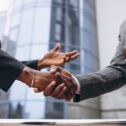 Two business professionals in formal suits shaking hands outdoors in front of a modern office building, symbolizing partnership and collaboration in infrastructure or corporate projects.