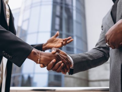 Two business professionals in formal suits shaking hands outdoors in front of a modern office building, symbolizing partnership and collaboration in infrastructure or corporate projects.