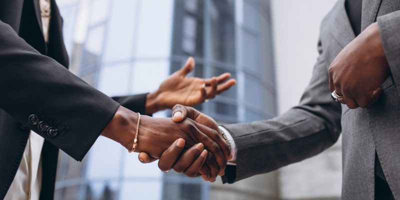 Two business professionals in formal suits shaking hands outdoors in front of a modern office building, symbolizing partnership and collaboration in infrastructure or corporate projects.