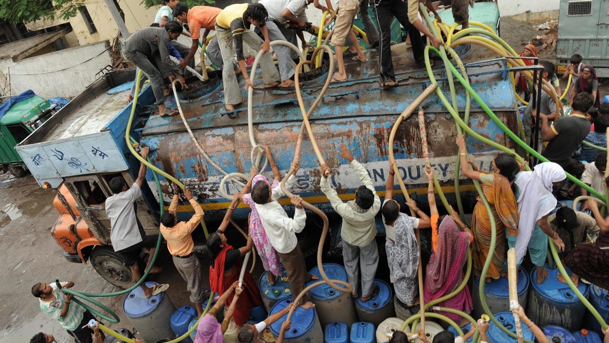 Crowd of people collecting water from a tanker truck using hoses and containers in an urban Indian area, highlighting water scarcity and infrastructure challenges.