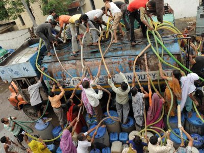 Crowd of people collecting water from a tanker truck using hoses and containers in an urban Indian area, highlighting water scarcity and infrastructure challenges.