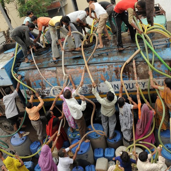 Crowd of people collecting water from a tanker truck using hoses and containers in an urban Indian area, highlighting water scarcity and infrastructure challenges.