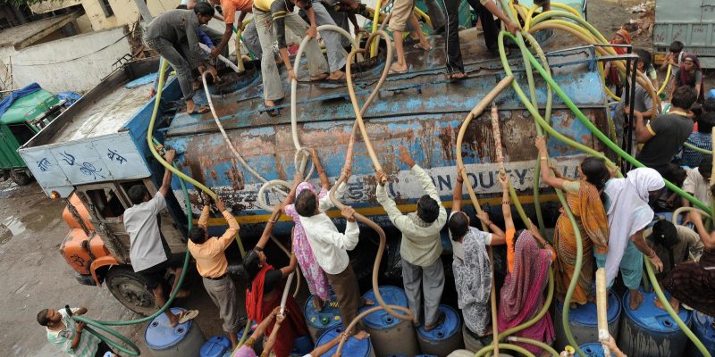 Crowd of people collecting water from a tanker truck using hoses and containers in an urban Indian area, highlighting water scarcity and infrastructure challenges.