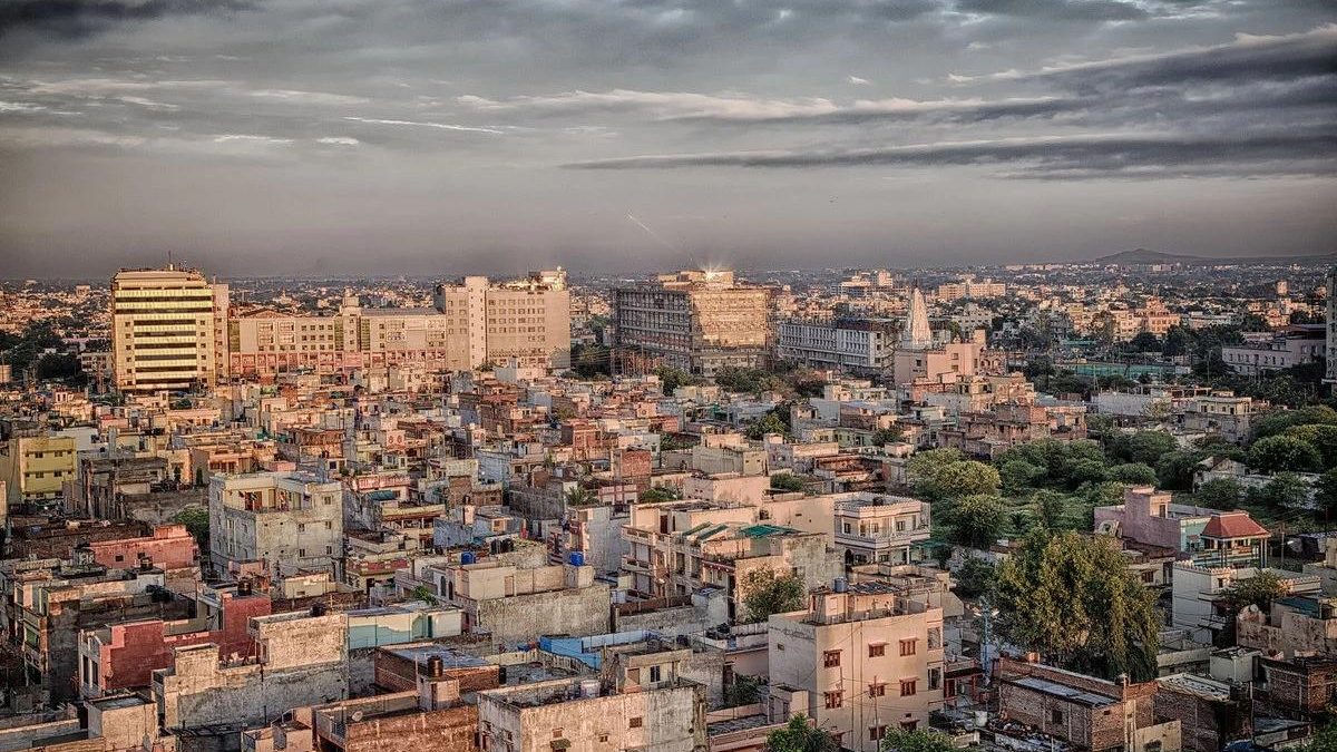 Wide view of a Tier-2 Indian city with dense low-rise housing, mid-rise commercial buildings, and a growing urban skyline under a cloudy sky.