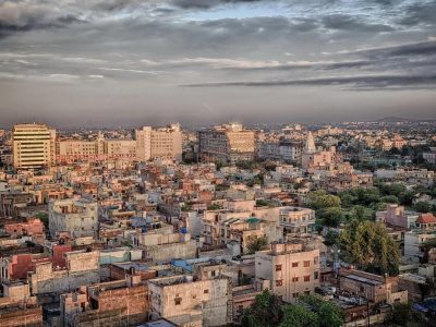 Wide view of a Tier-2 Indian city with dense low-rise housing, mid-rise commercial buildings, and a growing urban skyline under a cloudy sky.