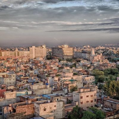 Wide view of a Tier-2 Indian city with dense low-rise housing, mid-rise commercial buildings, and a growing urban skyline under a cloudy sky.