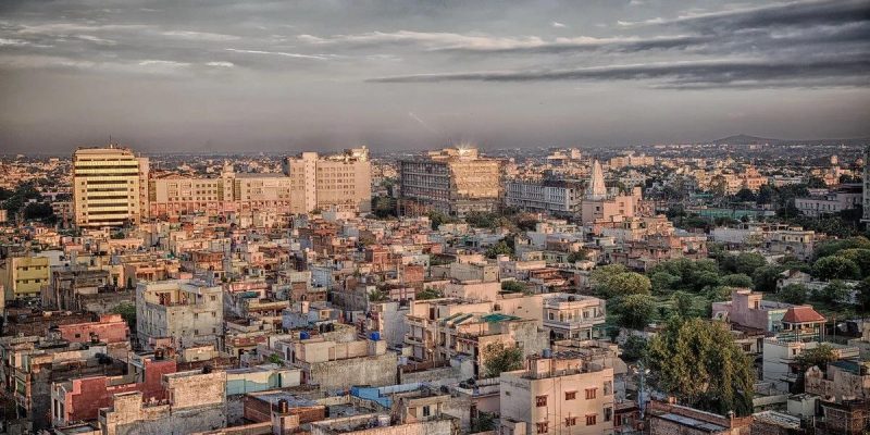 Wide view of a Tier-2 Indian city with dense low-rise housing, mid-rise commercial buildings, and a growing urban skyline under a cloudy sky.