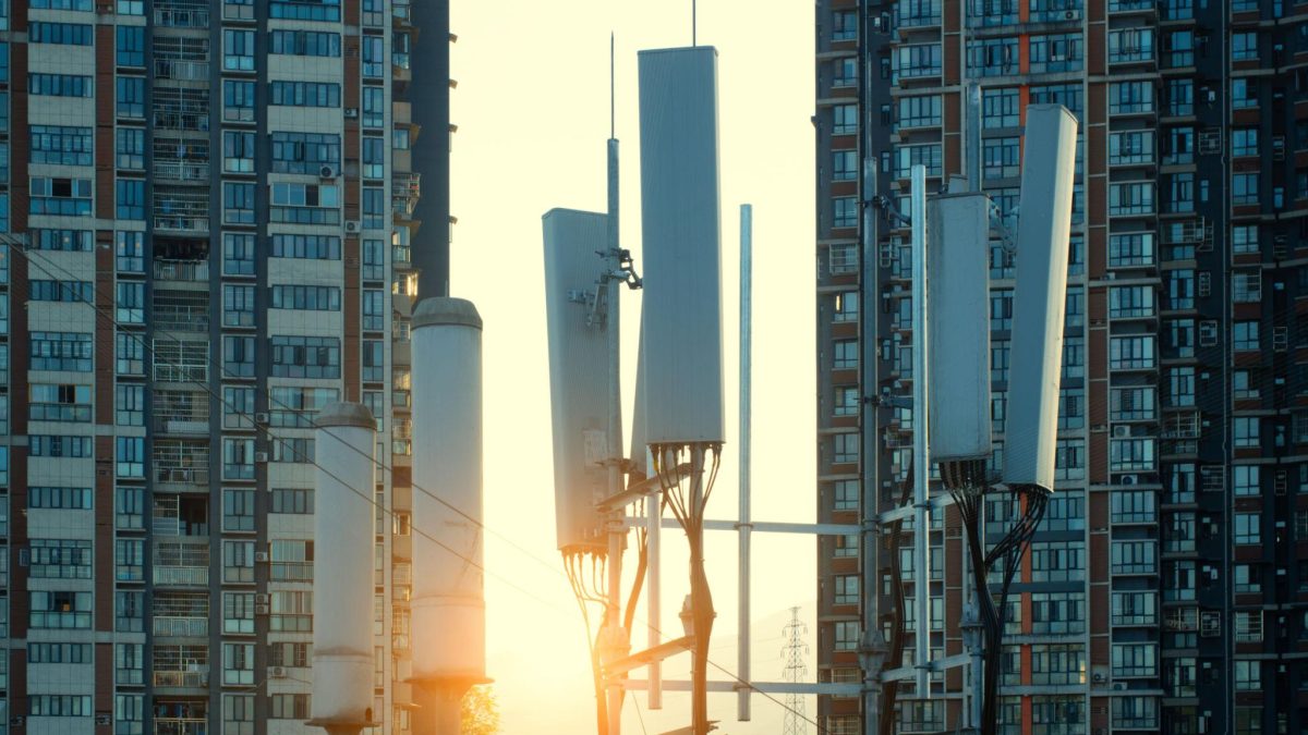 5G cellular antennas mounted on a telecom tower between high-rise residential buildings at sunset, representing urban digital infrastructure and connectivity.