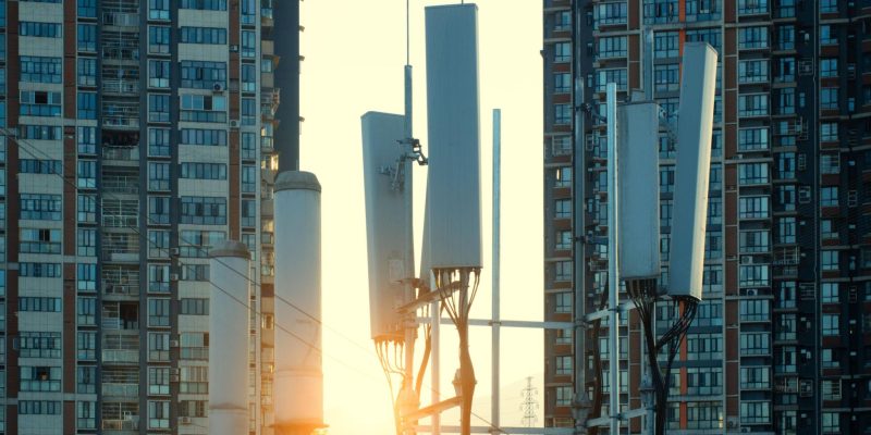 5G cellular antennas mounted on a telecom tower between high-rise residential buildings at sunset, representing urban digital infrastructure and connectivity.
