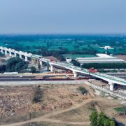 Aerial view of an elevated high-speed rail corridor under construction crossing a highway in a semi-rural landscape, with concrete pillars, construction equipment, and surrounding fields visible.