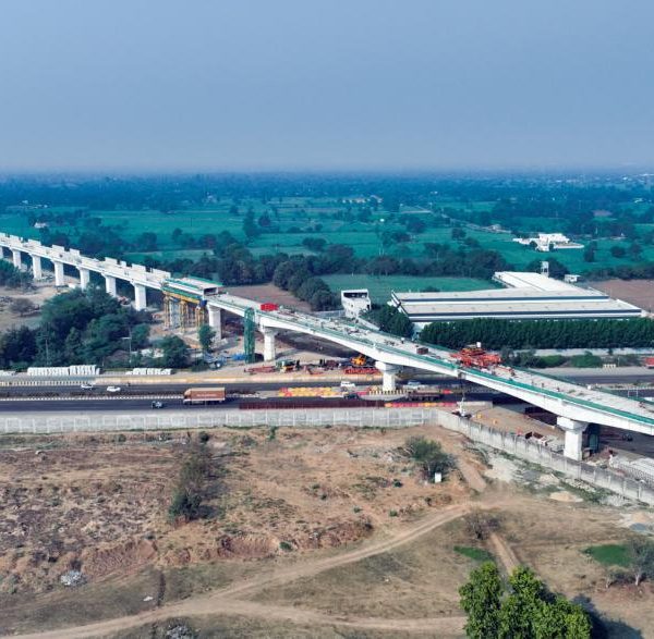 Aerial view of an elevated high-speed rail corridor under construction crossing a highway in a semi-rural landscape, with concrete pillars, construction equipment, and surrounding fields visible.