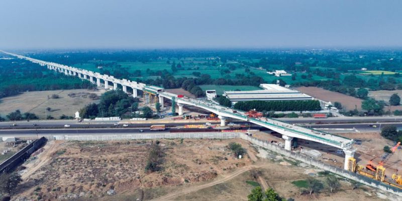 Aerial view of an elevated high-speed rail corridor under construction crossing a highway in a semi-rural landscape, with concrete pillars, construction equipment, and surrounding fields visible.
