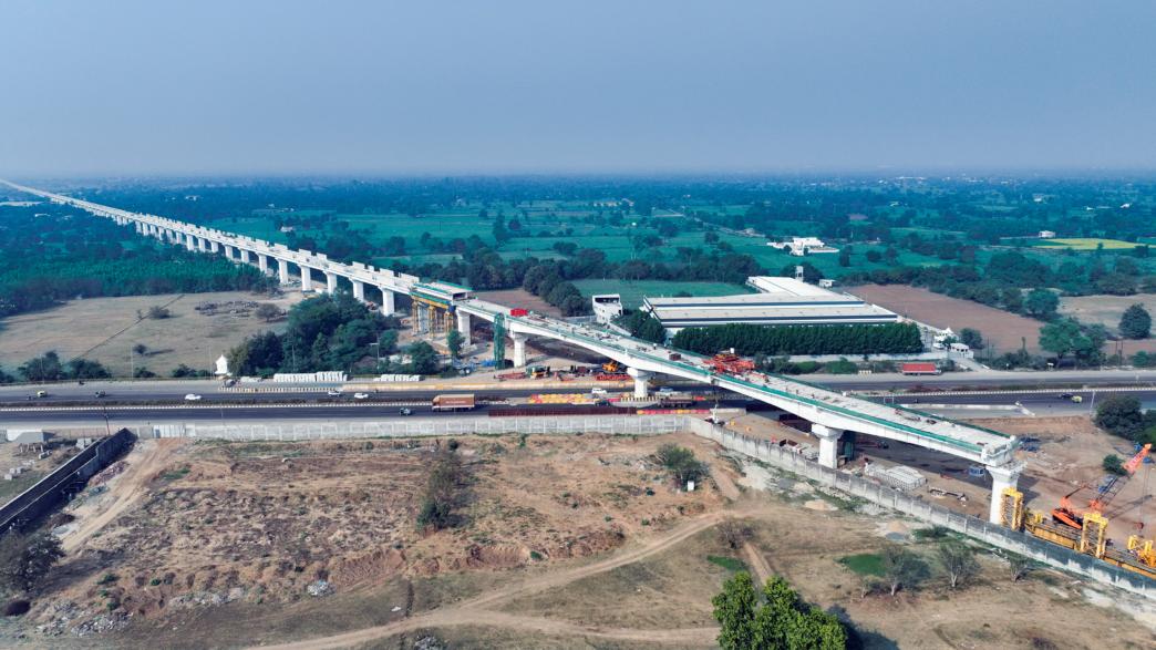 Aerial view of an elevated high-speed rail corridor under construction crossing a highway in a semi-rural landscape, with concrete pillars, construction equipment, and surrounding fields visible.