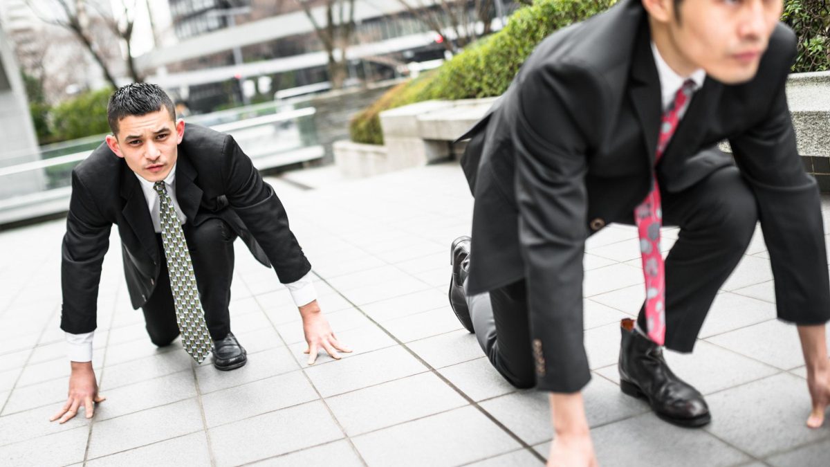 Two businessmen in suits crouched in a starting position on a city sidewalk, symbolizing competition and a race to gain advantage in business.