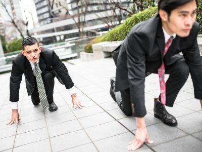 Two businessmen in suits crouched in a starting position on a city sidewalk, symbolizing competition and a race to gain advantage in business.