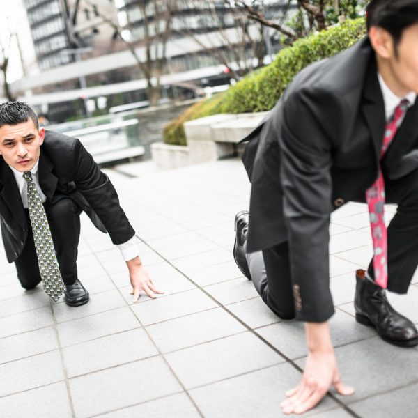 Two businessmen in suits crouched in a starting position on a city sidewalk, symbolizing competition and a race to gain advantage in business.
