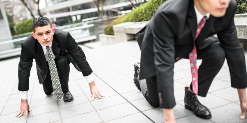 Two businessmen in suits crouched in a starting position on a city sidewalk, symbolizing competition and a race to gain advantage in business.