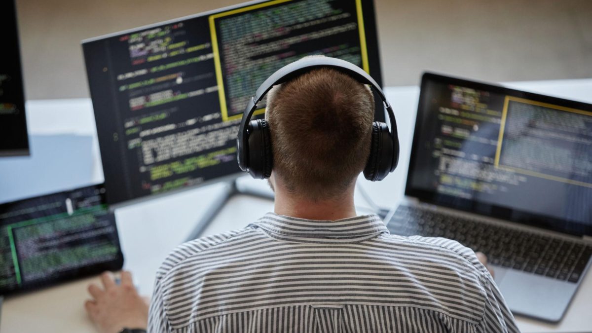 Developer wearing headphones working on multiple screens displaying code on a laptop and monitors in a modern workspace.
