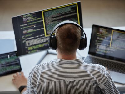 Developer wearing headphones working on multiple screens displaying code on a laptop and monitors in a modern workspace.