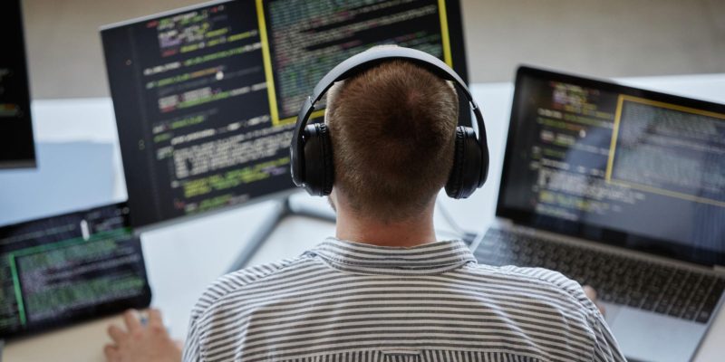 Developer wearing headphones working on multiple screens displaying code on a laptop and monitors in a modern workspace.