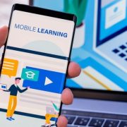 Person holding a smartphone displaying a mobile learning app, with a laptop in the background.