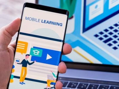 Person holding a smartphone displaying a mobile learning app, with a laptop in the background.