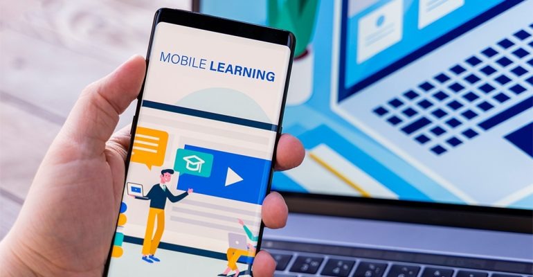 Person holding a smartphone displaying a mobile learning app, with a laptop in the background.