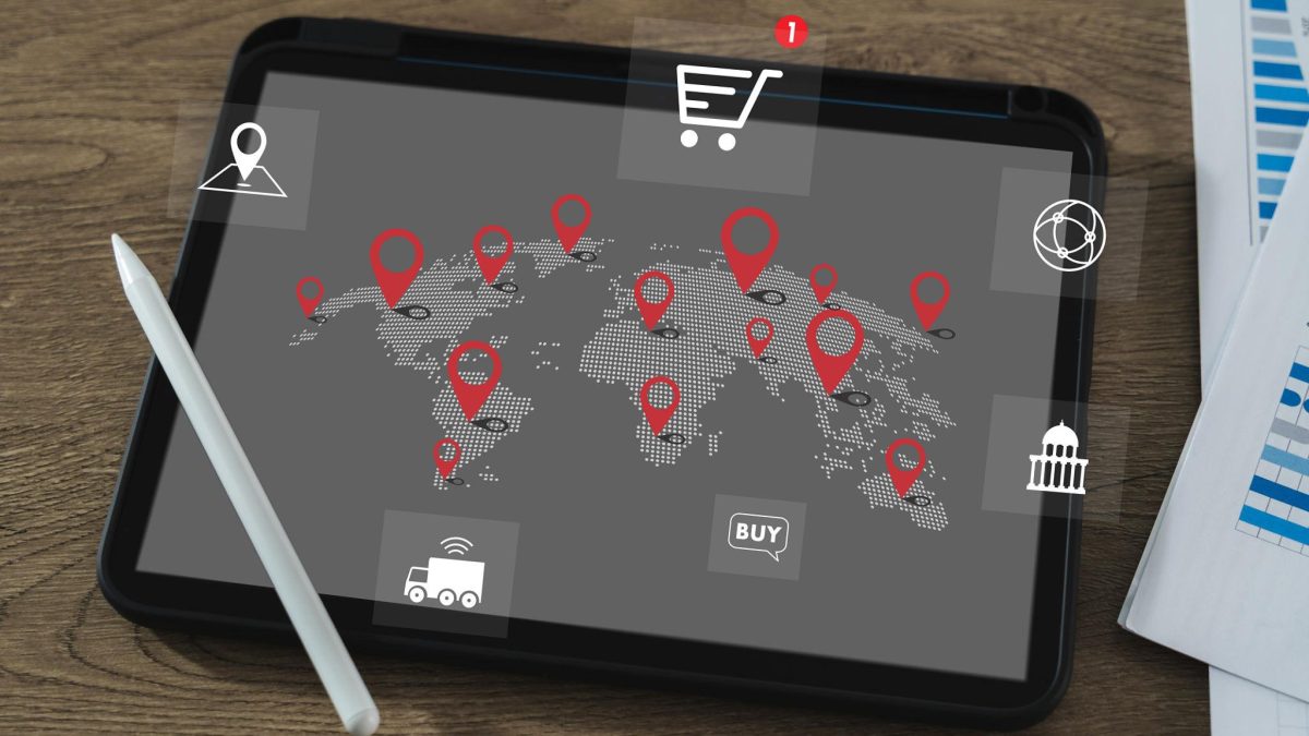 Tablet displaying a world map with location pins and global trade icons, placed on a desk with a stylus and documents.