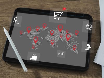 Tablet displaying a world map with location pins and global trade icons, placed on a desk with a stylus and documents.