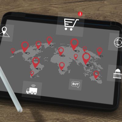 Tablet displaying a world map with location pins and global trade icons, placed on a desk with a stylus and documents.