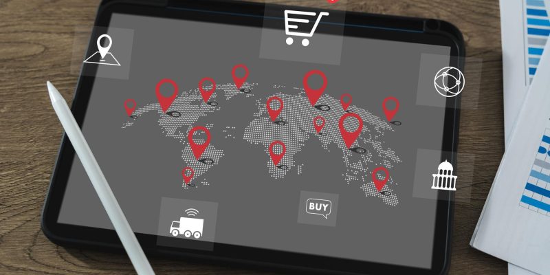Tablet displaying a world map with location pins and global trade icons, placed on a desk with a stylus and documents.